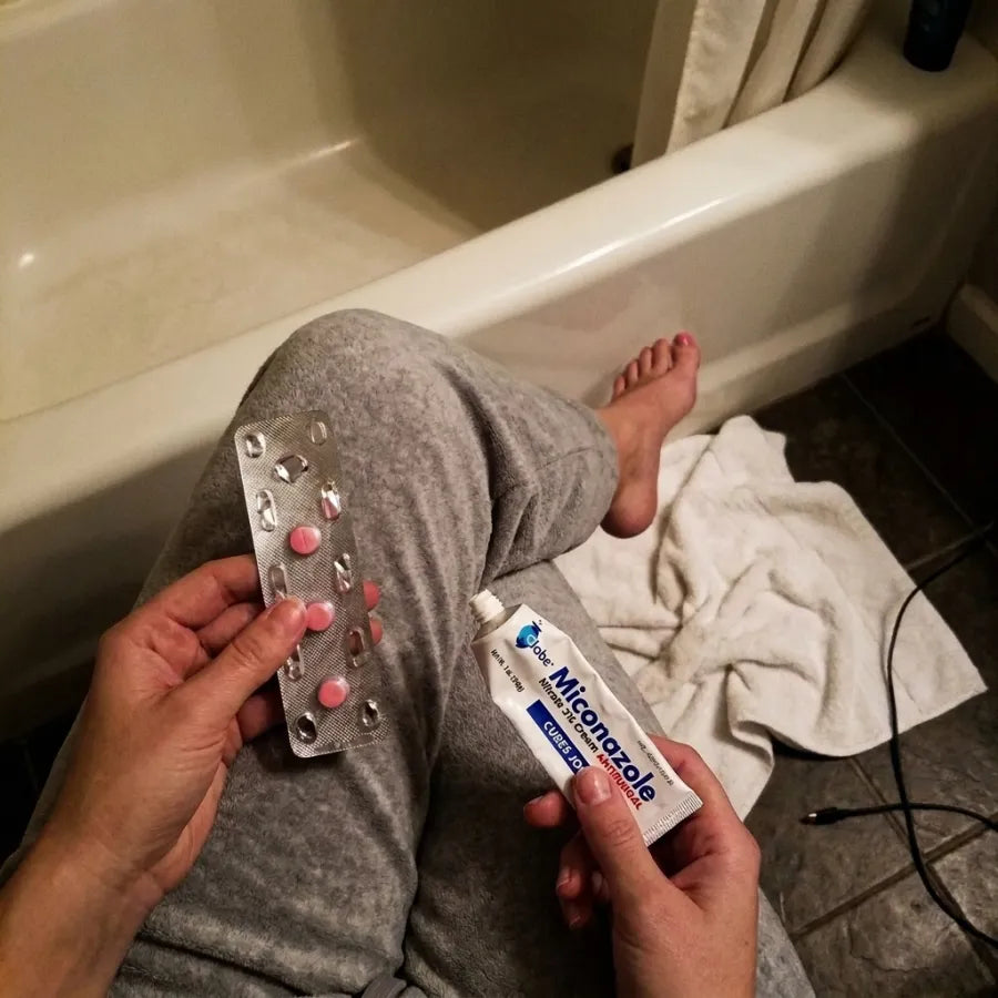 A woman sitting on the edge of her bathtub holding antifungal pills and miconazole cream.